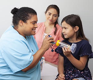 Healthcare provider showing girl how to use metered-dose inhaler with spacer and mask. Woman standing nearby.